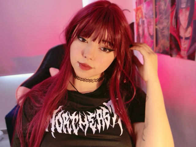 Arieyaara, con lunghi capelli rossi, un collare nero e una maglietta nera con la scritta "Gothic", guarda la fotocamera con un leggero sorriso. Illuminazione rosa e rossa, mano tra i capelli, tatuaggio sul braccio. Incredibilmente sexy..