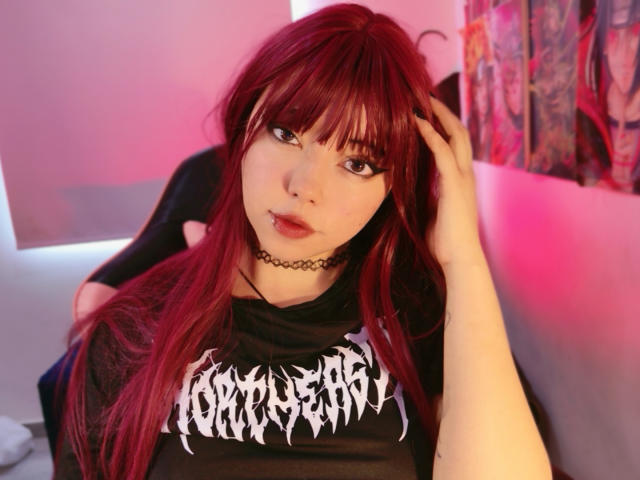 Photograph of Arieyaara with long, red hair, black choker, and black "Cobalt" t-shirt. She's touching her hair, lips slightly parted, pink and red lighting in the background. Fucking sexy.