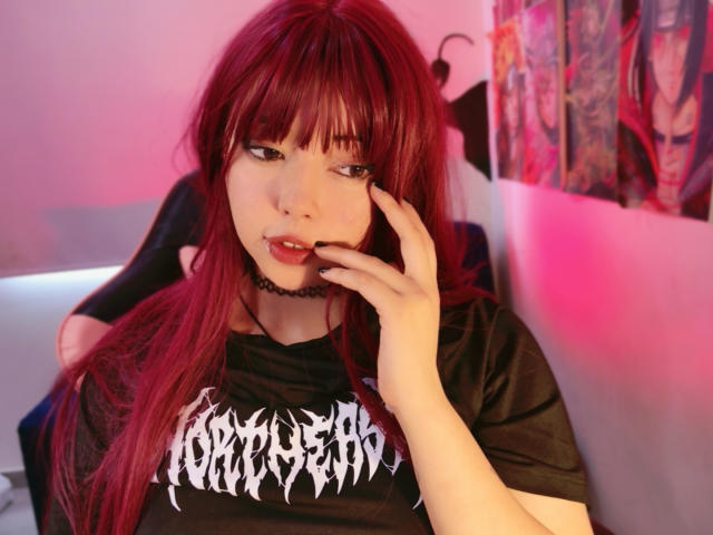 Arieyaara, mit roten Haaren, Pony und heller Haut, trägt ein schwarzes T-Shirt mit der Aufschrift "Crimson" und berührt ihre Lippen mit ihrer rechten Hand. Rosa und violette Beleuchtung erzeugt eine sinnliche, provokante Atmosphäre..