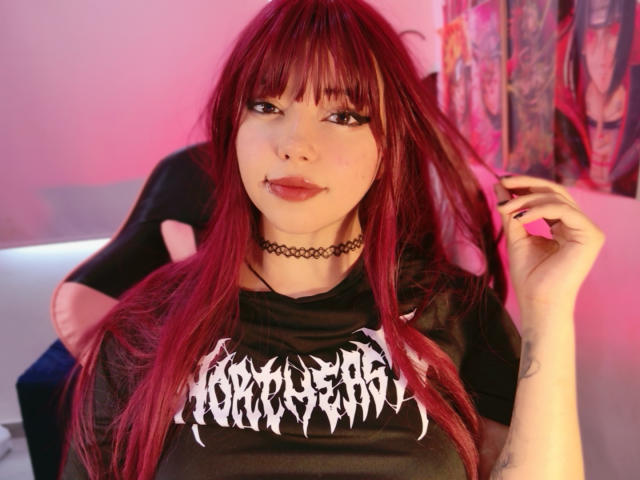 Foto van Arieyaara met lang, rood haar, een zwarte choker en een zwart "Iron Maiden" T-shirt. Ze houdt haar haar vast en likt haar lip, met een verleidelijke blik. Roze en rode verlichting.