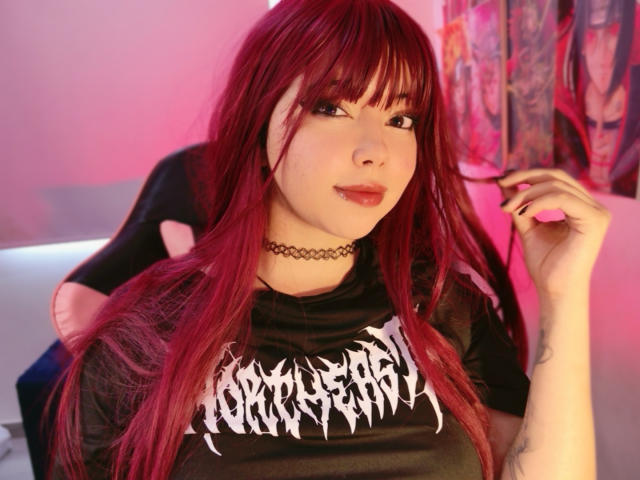 Arieyaara, con lunghi capelli rossi, un collare nero e una maglietta nera con la scritta "Berserk", seduta con un braccio di un ragazzo intorno al collo, indossa qualcosa di rosa. e luci rosse, tatuaggi sul braccio, espressione seducente.