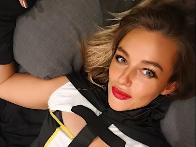 Primer plano de RebeccaNoelle acostada sobre un cojín gris, vistiendo una camiseta blanco y negro con una franja amarilla, mirando directamente. mirando a la cámara con un lápiz labial rojo brillante, ojos azules y cabello rubio..