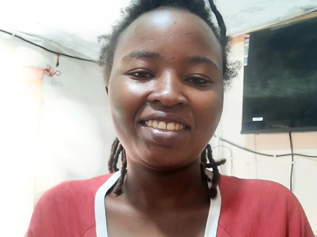 Close-up of RachieChristina with dark skin, smiling, wearing a red shirt with a white collar, and braided hair. Background is white with black and beige elements. Slight sheen on face.