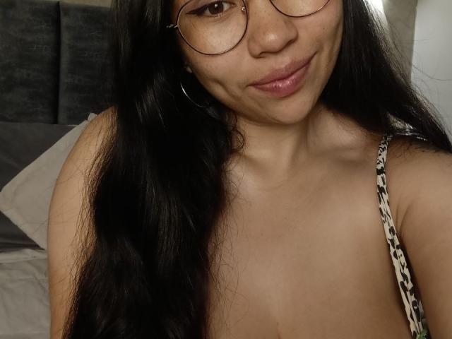 Gros plan de Karollteyes, avec de longs cheveux noirs, des lunettes rondes et un léger sourire. Elle est nue sur le buste, laissant entrevoir une partie de ses seins avec une bretelle ornée qui traverse une de ses épaules. Extrêmement séduisant..