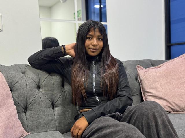 NahomyNoir with long, straight black hair sits on a gray tufted couch, wearing a black zip-up top and gray pants, hand behind head, slight smile, pink pillows, sexual tension.