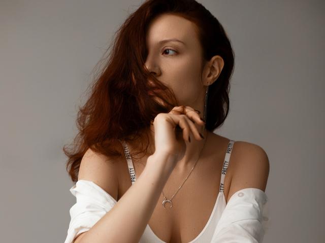 BarbaraStily heeft lang, golvend bruin haar dat haar rechteroog bedekt en kijkt naar links. Ze draagt een witte top met een open schouderlijn en smalle bandjes, en een delicate ketting. Heel aantrekkelijk..