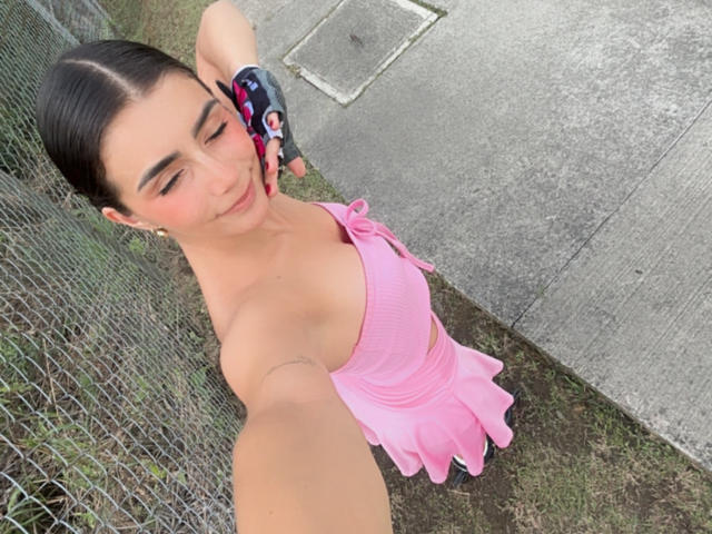 KimmyK prend un selfie d'en haut, montrant sa peau bronzée et ses cheveux noirs attachés en chignon. Elle porte une robe rose, ornée de volants, qui révèle ses petits seins. L'arrière-plan comprend une clôture en treillis métallique et un trottoir..