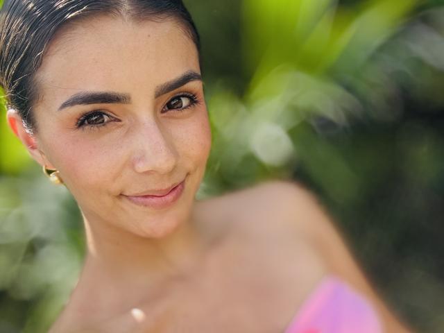 Close-up of KimmyK, a sexy woman with dark hair pulled back, brown eyes, and a subtle smile. She's wearing a pink strapless top, with greenery blurred in the background.