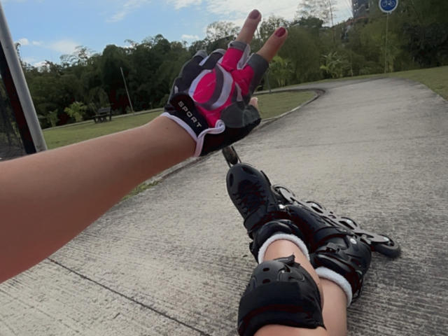 Photograph of a KimmyK lying on a concrete path, wearing black rollerblades and black gloves with pink accents, making a peace sign with the right hand, outdoor setting.