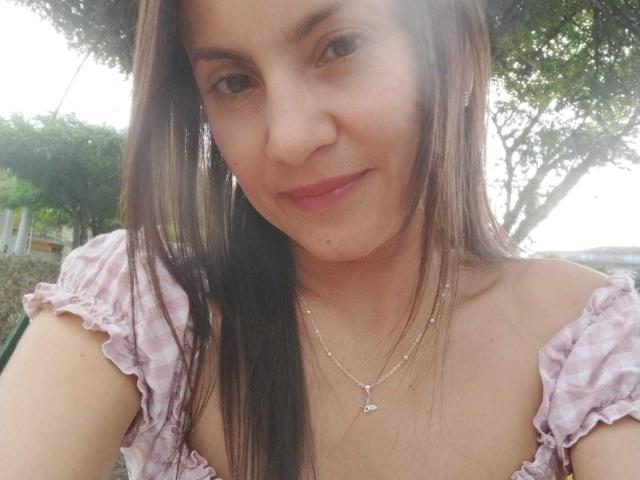 Close-up selfie of SolaraDubois, a young woman with straight brown hair, light skin, and a subtle smile. She wears a pink and white checkered top, visible cleavage, and a delicate necklace. Fucking hot.