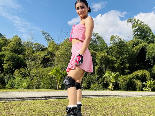 KimmyK está ao ar livre, em um gramado, vestindo um vestido rosa sem alças, joelheiras pretas e tênis pretos. Ela está sorrindo com uma postura confiante, demonstrando um leve toque de tensão sexual. Céu azul brilhante.