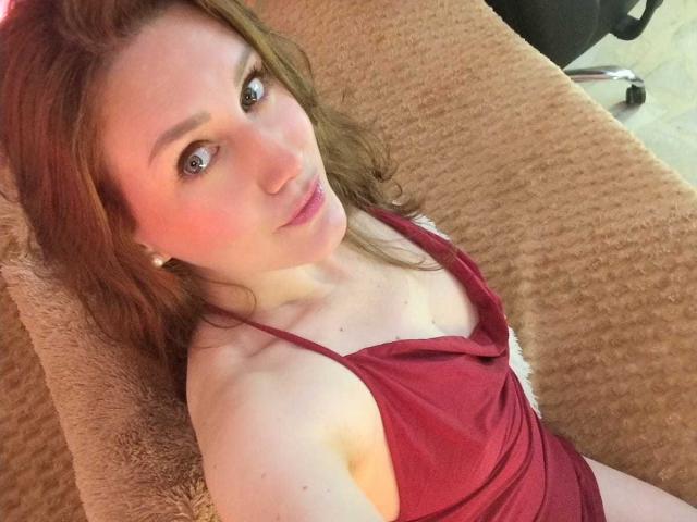 Photograph of KatyaRogersX with long brown hair, lying on a brown textured couch, wearing a red tank top, looking up with a slightly open mouth, light skin, relaxed pose, sexual tension.