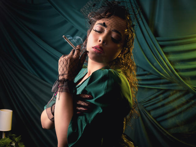 Barbara Leythonn, avec ses longs cheveux noirs, les yeux fermés, tenant une cigarette dans des gants en dentelle noire, portant une robe verte, penchée en arrière, fond avec des rideaux sombres, éclairage tamisé, expression sexy et aguicheuse..