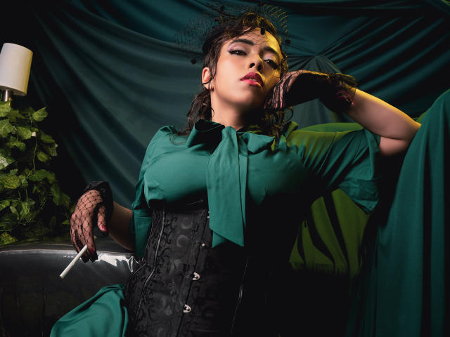 BarbaraLeythonn sits in a dark, moody setting, wearing a green shirt and black corset, with black lace gloves holding a cigarette. Her hair is messy, expression sultry. Lighting is dramatic.