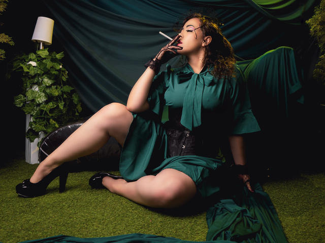 Barbara Leythonn est assise sur l'herbe verte, vêtue d'une robe verte, d'un corset noir et de gants noirs, et elle fume une cigarette. Elle a les cheveux bruns bouclés, des lunettes et des talons hauts noirs. Une ambiance sombre et sensuelle..