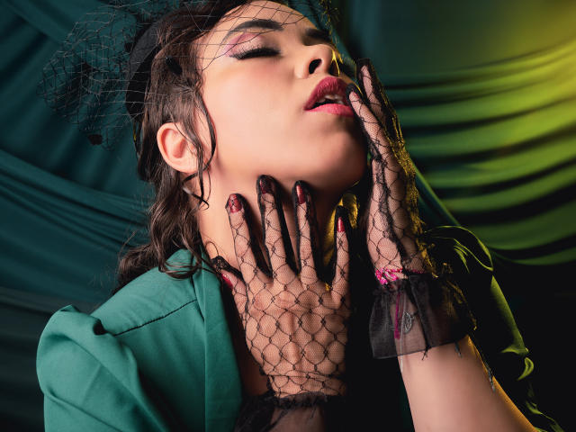 Barbara Leythonn, occhi socchiusi, labbra leggermente aperte, indossa guanti di pizzo nero, una giacca verde, capelli scuri, un velo nero, chiara di carnagione. Sfondo verde-giallo, mano al collo, rossetto rosso, posa seducente, espressione intensa, texture lucida..