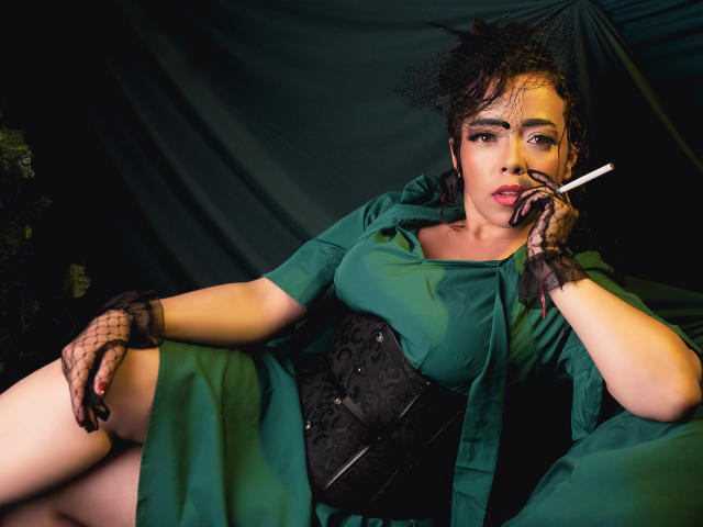 BarbaraLeythonn lounges seductively in a green dress, black corset, and fishnet gloves, holding a cigarette. Her dark curly hair, intense gaze, and slightly parted lips create intense sexual tension.