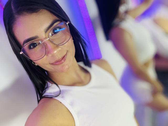 Close-up of KimmyK, a brunette woman with blue eyes, wearing clear glasses and a white sleeveless top. Blurred background with another person, hint of sexual tension. Vibrant colors.