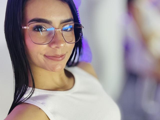 Close-up van KimmyK met recht zwart haar, die heldere brillen draagt die blauw licht reflecteren, en een wit topje zonder mouwen. Ze heeft een subtiele grijns, haar huid is glad, en er is een vage figuur op de achtergrond..