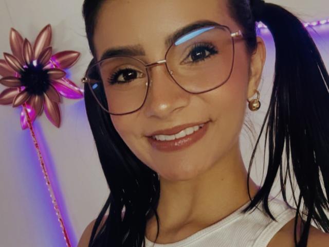 Close-up of KimmyK with dark pigtails, wearing large round glasses, white tank top, and gold earrings. Pink flower-shaped toy with neon lights on left. Smiling, showing white teeth.