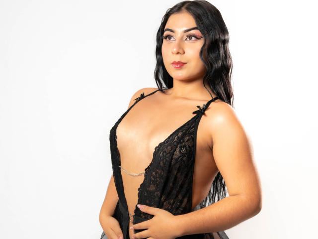 Gaby Taylor, mit langen, schwarzen Haaren, trägt ein tief ausgeschnittenes, schwarzes Spitzenkleid, die Hände an den Hüften, wobei ihr Dekolleté zu sehen ist, weiß. Hintergrund, direkter Blick, volle Lippen, leicht gewölbte Augenbrauen, sexy, selbstbewusste Pose.