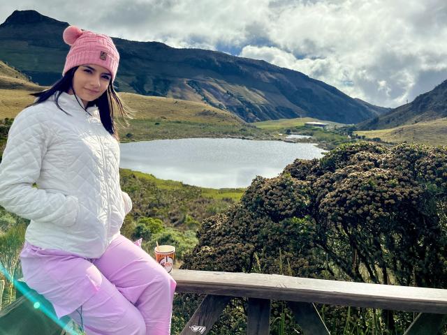 KimmyK with dark hair, pink beanie, white quilted jacket, and pink pants, leaning on a wooden railing, mountains and lake in the background, bright sunlight, sexual tension, casual pose.