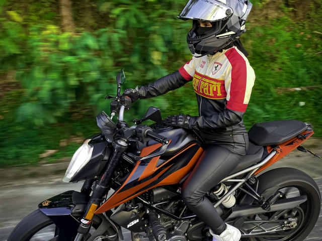 KimmyK on a black and orange motorcycle, wearing a white helmet, red and yellow shirt, black gloves, and black pants. Blurred green background, riding forward, cocked head, intense focus.