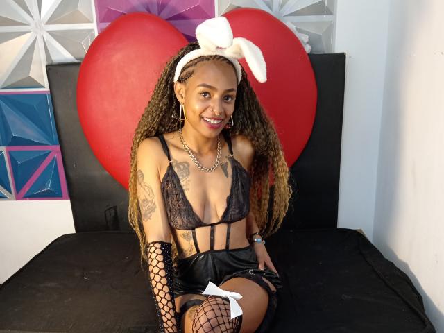 KatalinaCandy, avec ses longs cheveux bouclés, portant un soutien-gorge en dentelle noire, des gants en résille, un short noir et un serre-tête en forme de lapin blanc, assise sur un lit noir, avec un cœur rouge derrière elle, souriante..