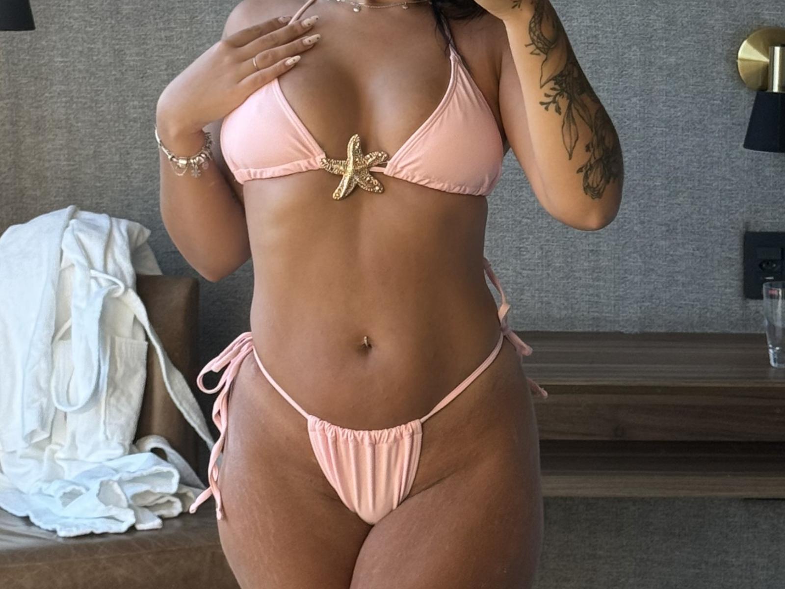 BelssBarney portant un bikini de couleur pêche avec un nœud doré, une main couvrant sa poitrine, un bras tatoué, une silhouette voluptueuse, un léger bronzage. Debout dans une pièce faiblement éclairée, un tissu blanc sur la table. Tension sexuelle.