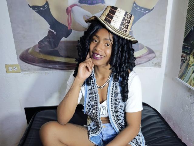 KatalinaCandy sitzt auf einem schwarzen Ledersofa und lächelt, mit einem Finger am Mund. Sie hat langes, schwarzes Haar, trägt ein weißes Hemd und eine blaue Jeansshorts sowie einen Strohhut. Verdammt heiß.
