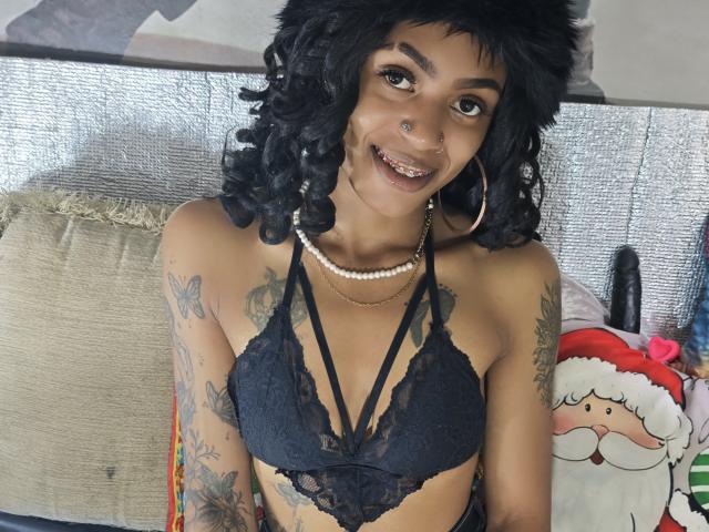 KatalinaCandy mit kurzen, lockigen, schwarzen Haaren, dunkler Haut und Tätowierungen, trägt ein schwarzes Spitzen-Bralette und eine Perlenkette. mit einem Nasenring lächelnd. Weihnachtsmann-Plüschtier rechts. Verdammt sexy..