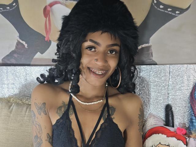 Fotografie von KatalinaCandy, die lächelnd mit schwarzen, lockigen Haaren zu sehen ist, und die ein schwarzes Spitzen-Haltertop, goldene Ohrringe im Stecker-Design und perlenkette. Tattoos an den Armen. Im Hintergrund ist ein sexy Poster und Spielzeug zu sehen..
