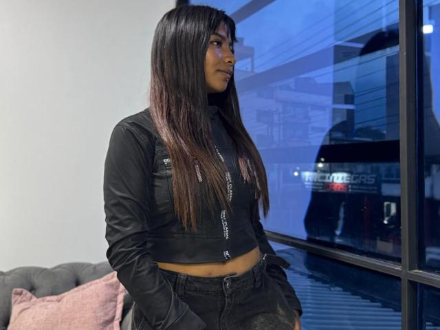 Photograph of NahomyNoir with long, straight black hair, wearing a black crop top and high-waisted jeans, standing by a window at night. Reflection of a truck in the glass. Blue lighting.