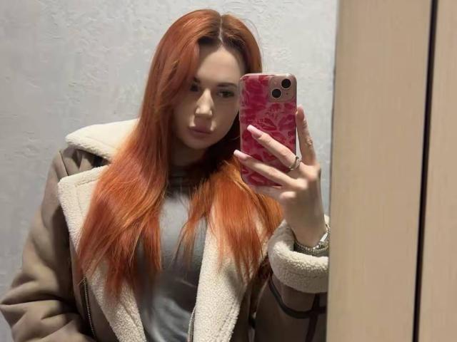Fotografía de JuicyJoyce tomando una selfie en el espejo. Ella tiene el pelo largo y rojo, lleva un abrigo marrón con un cuello de piel blanca, sostiene un teléfono rosa y mira a la cámara. expresión seria.