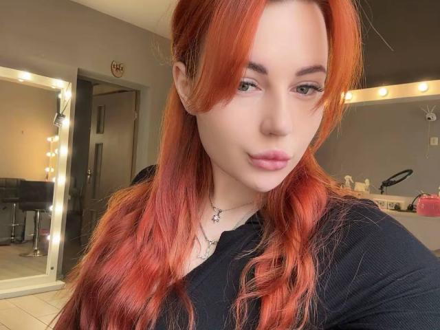 Close-up of JuicyJoyce with red hair, wearing a black top, looking directly at the camera with a slight smile. Soft lighting from a mirror in the background. Sexy, relaxed vibe.