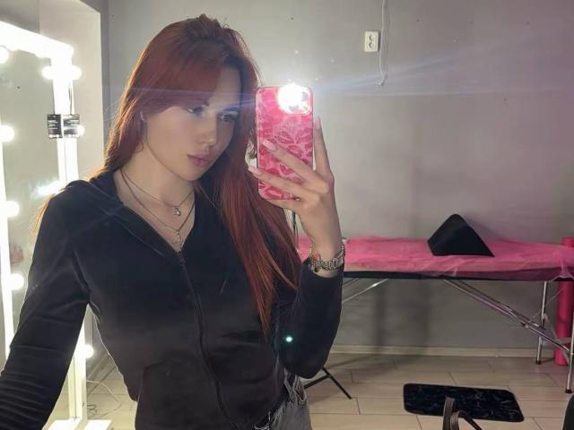 Fotografie von JuicyJoyce, die ein Selfie vor einem Spiegel macht. Sie hat langes, rotes Haar, trägt eine schwarze Kapuzenjacke und hält ein pinkes Handy mit einer glitzernden Hülle. Sie steht in der Nähe einer beleuchteten Wand. und eine pinke Bank. Ihr Ausdruck ist neutral, mit einem leichten Hauch von Verführung. Das helle Licht spiegelt sich auf dem Bildschirm ihres Handys wider..