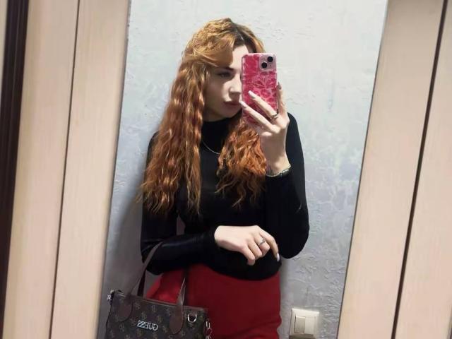 JuicyJoyce, met lang, golvend rood haar, maakt een selfie in de spiegel met een roze telefoon, en draagt een zwarte top en een rode rok. met een zwarte handtas, staand voor een witte muur.