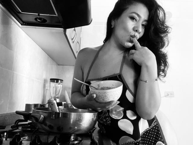 Black-and-white photo of AlisGourmande with long hair, holding a bowl, leaning on a stovetop, wearing a low-cut, polka-dot apron, biting her finger, with a stove and blender in the background.