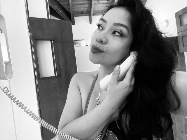 Fotografia em preto e branco de AlisGourmande, uma mulher atraente com cabelos longos e escuros, segurando um telefone com fio no ouvido. Fazendo um leve beicinho, vestindo um top sem alças, com uma forte tensão sexual..