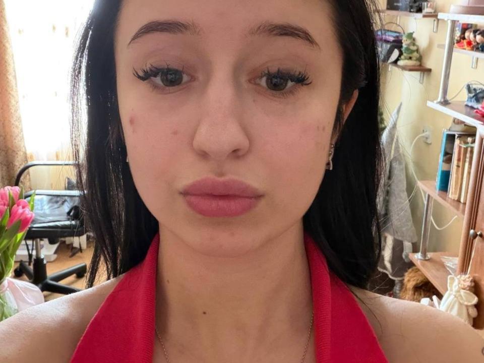 Fotografia ravvicinata di WinnieSemaan, una donna dalla pelle chiara e con lunghi capelli neri, che indossa un top rosso a spalline e guarda direttamente verso l'obiettivo. la fotocamera con labbra piene e ciglia scure e folte. Lo sfondo mostra una stanza disordinata..