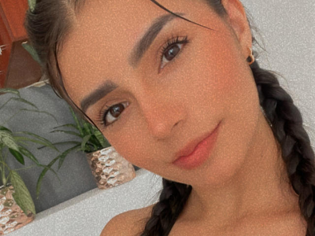 Close-up of KimmyK, a young woman with olive skin, dark braided hair, and thick eyebrows. She has a subtle smile, dark eyes, and is wearing small earrings. Background includes a potted plant.