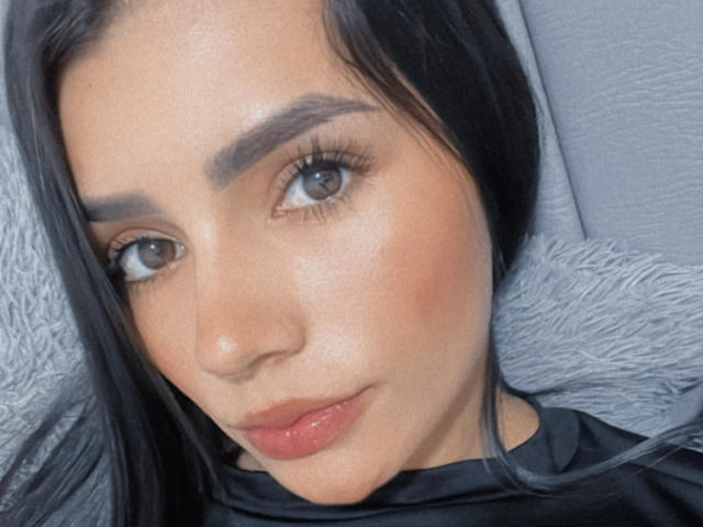 Close-up selfie of KimmyK, a woman with light brown skin, dark eyebrows, and black hair. She has full lips, a slight smirk, and brown eyes. Wearing a black top. Sexual tension evident.