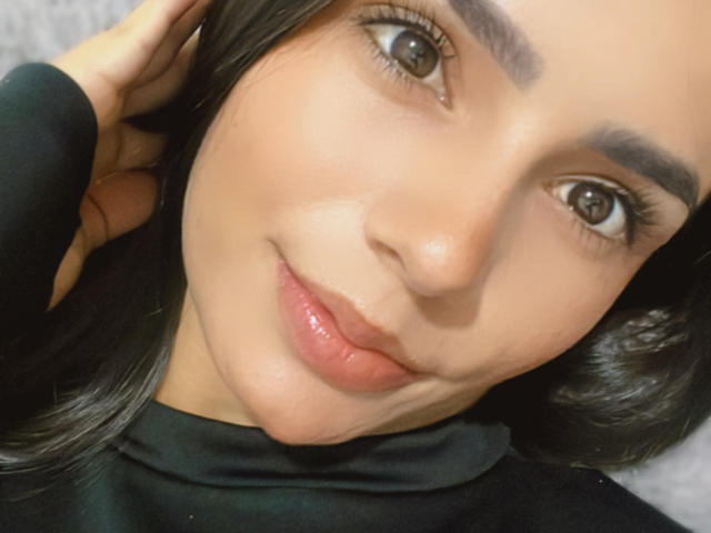 Close-up of KimmyK's face with smooth, light brown skin, full lips, and dark, thick eyebrows. KimmyK's large brown eyes gaze directly at the camera, wearing a black top. Fucking sexy.