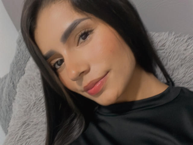 Primer plano de KimmyK, una mujer con cabello negro largo, cejas pobladas y labios rojos, vestida con una blusa negra, inclinándose ligeramente. a la izquierda, mirando directamente a la cámara con una sonrisa sutil.