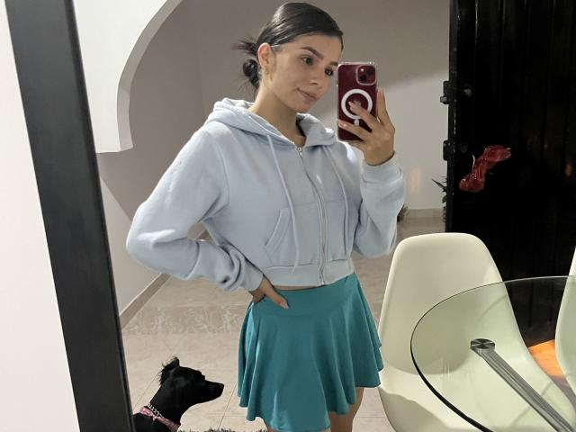 KimmyK staat voor een spiegel en maakt een selfie, terwijl ze een rode telefoon vasthoudt met een witte popsocket. Ze draagt een lichtblauwe hoodie en een turquoise rok, met haar hand op haar heup, en er ligt een zwarte hond op de vloer..
