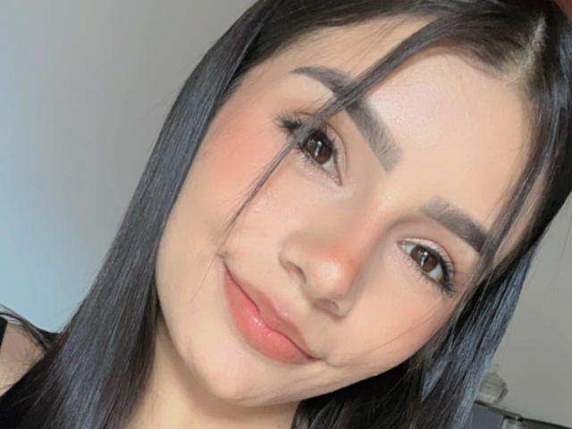 Close-up of KimmyK, a young woman with long black hair, fair skin, and brown eyes, smiling slightly. She has full eyebrows and subtle pink lipstick. Her expression is relaxed and slightly seductive.