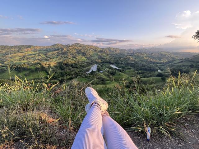 Photograph of KimmyK's legs extending into the foreground, wearing white, ribbed socks, with feet pointing towards a lush, mountainous landscape under a clear, blue sky at sunset. Slight sexual tension.
