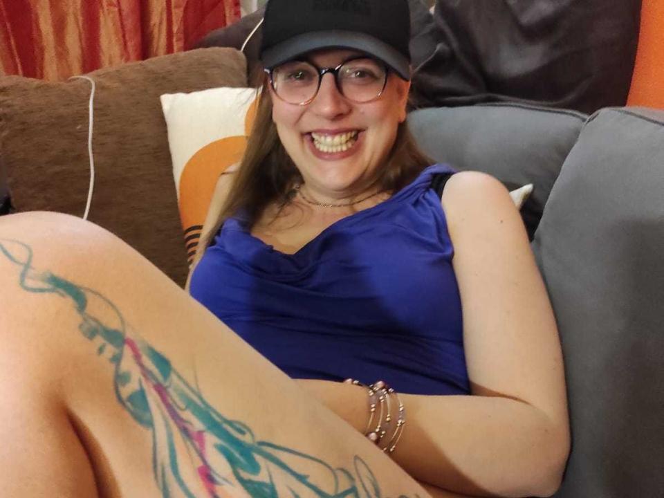 FlammeFrivole, wearing a black cap and glasses, sits on a couch with legs spread, showing a colorful thigh tattoo. She's smiling, wearing a blue dress, bracelet on wrist. Cock visible.