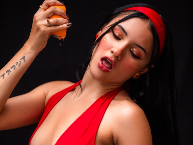 LizzieJoy, une femme séduisante avec des cheveux noirs et un bandeau rouge, porte une orange à ses lèvres, les yeux à moitié fermés, et un haut à enfiler rouge. montrant sa poitrine, tatouage sur l'avant-bras droit. Extrêmement excité(e)..