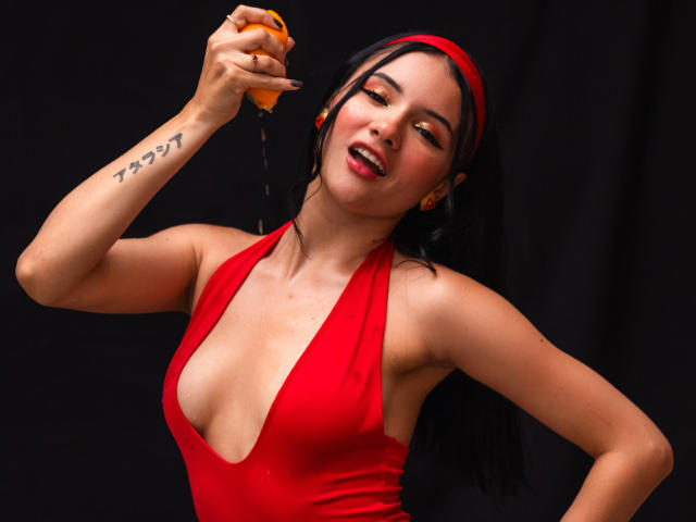 LizzieJoy, con i capelli scuri e una fascia rossa, indossa un abito rosso a scollo basso, tenendo in mano un'arancia da cui cola del liquido. Il tatuaggio sul suo braccio riporta il numero "7". Sta guardando seducentemente la fotocamera..